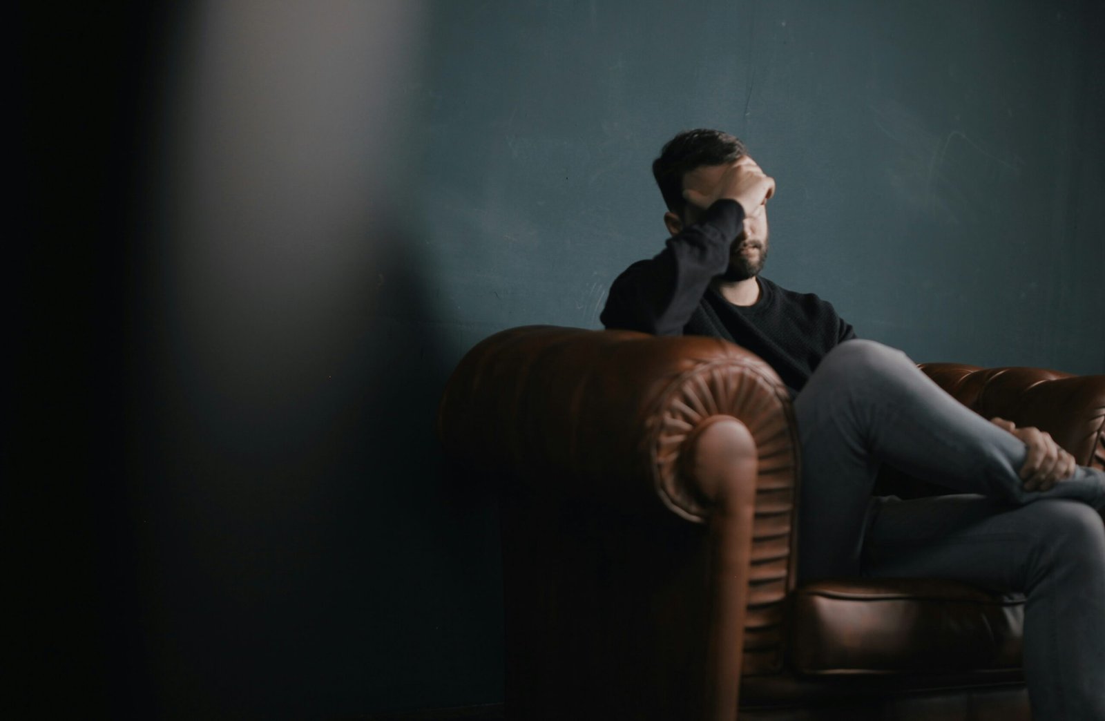 Man in black T‑shirt sitting on brown sofa, hand on forehead, symbolising fatigue linked to antibiotics and mental health