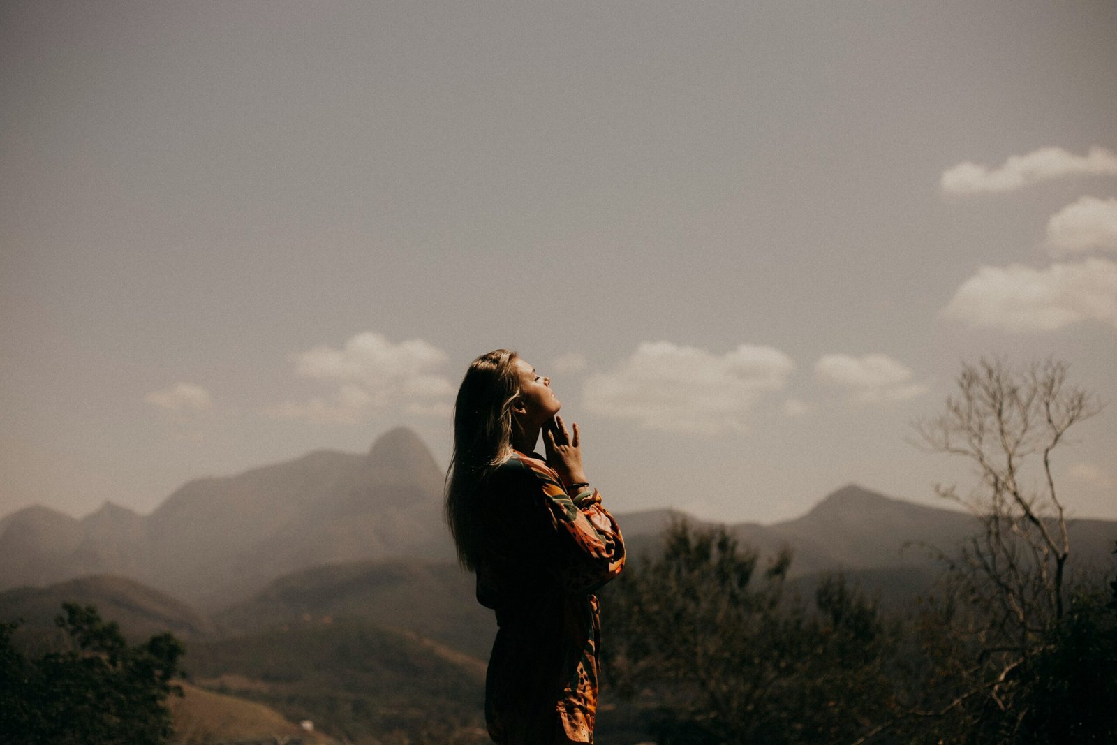 A woman standing in nature with her eyes closed, basking in sunlight, practicing mindfulness or deep breathing in a mountainous landscape.